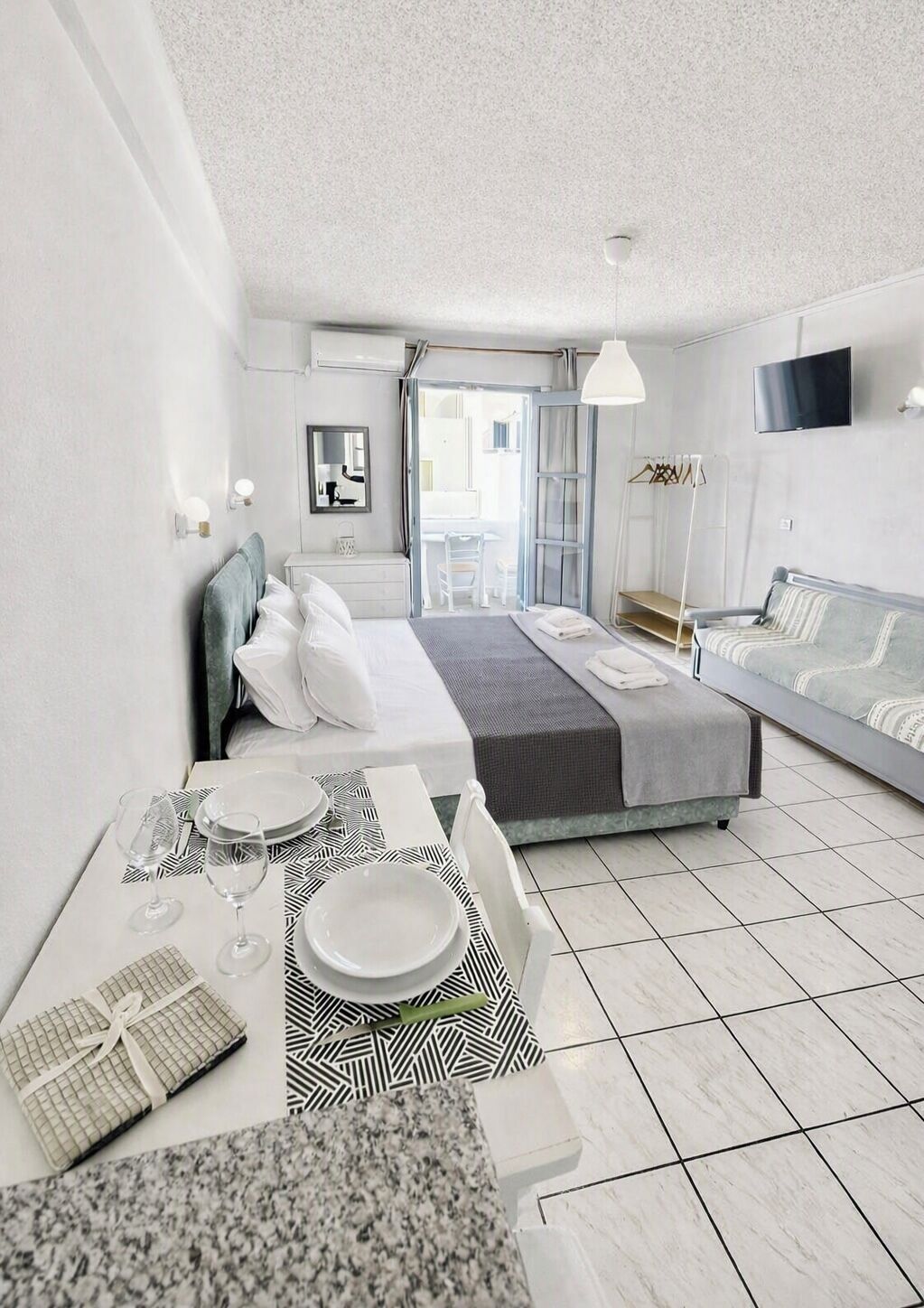 A modern hotel room with a bed, a sofa, a dining table with plates and glasses, and a television on the wall.