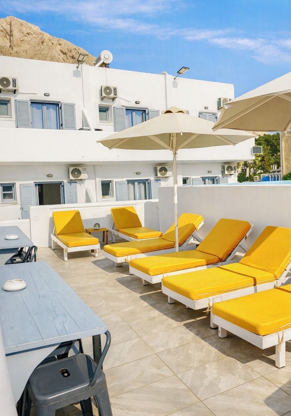 Un área de lounge en la azotea con tumbonas amarillas y sombrillas, unidades de aire acondicionado y vistas a un paisaje montañoso.