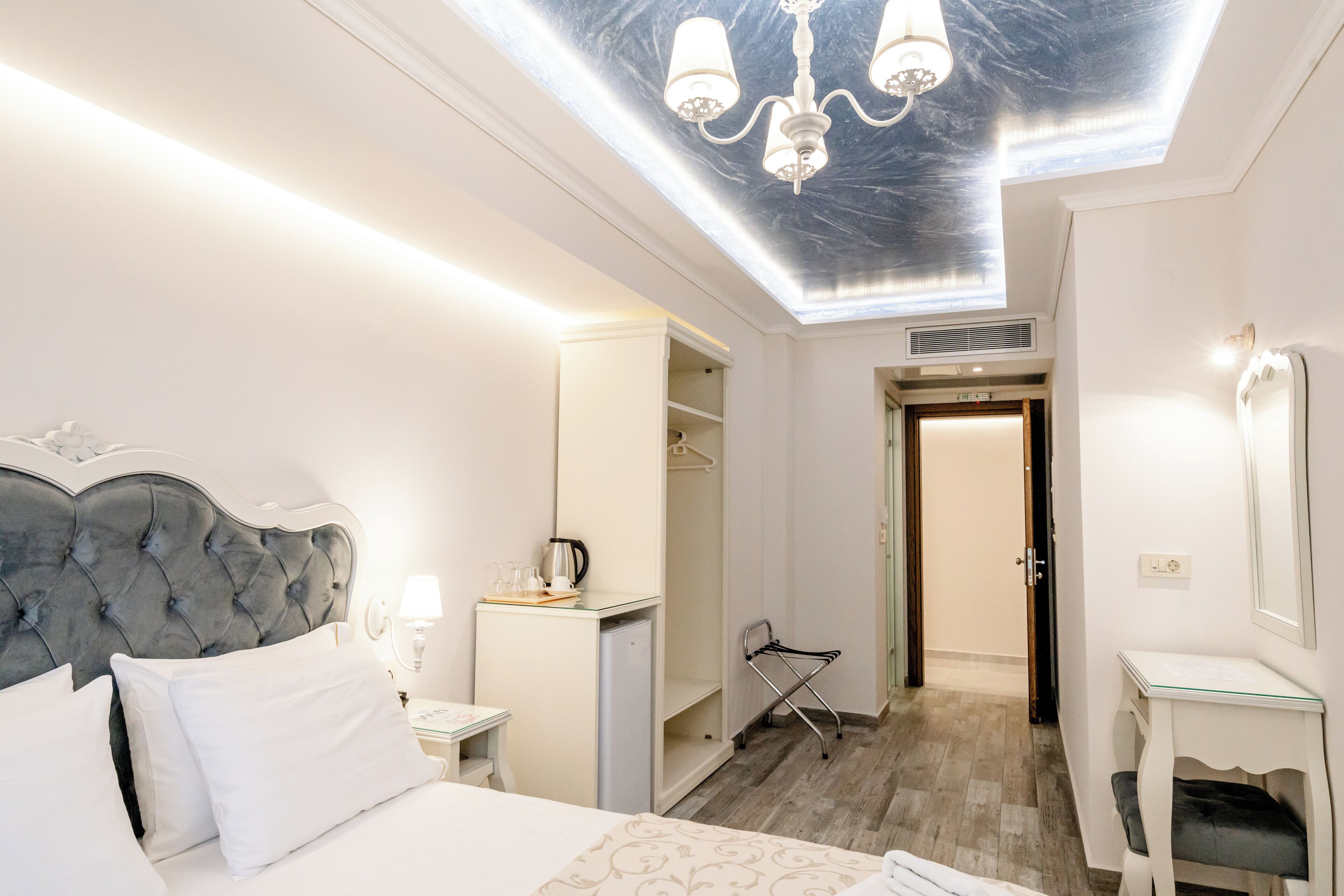 A hotel room with a tufted headboard, a bed with white linens, a bedside table with a lamp, a desk with a chair, a walk-in closet, and a skylight with a chandelier.