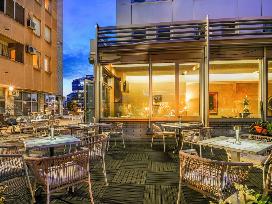 An outdoor dining area with wicker chairs and tables, a modern building facade, and a view of the interior through large windows.