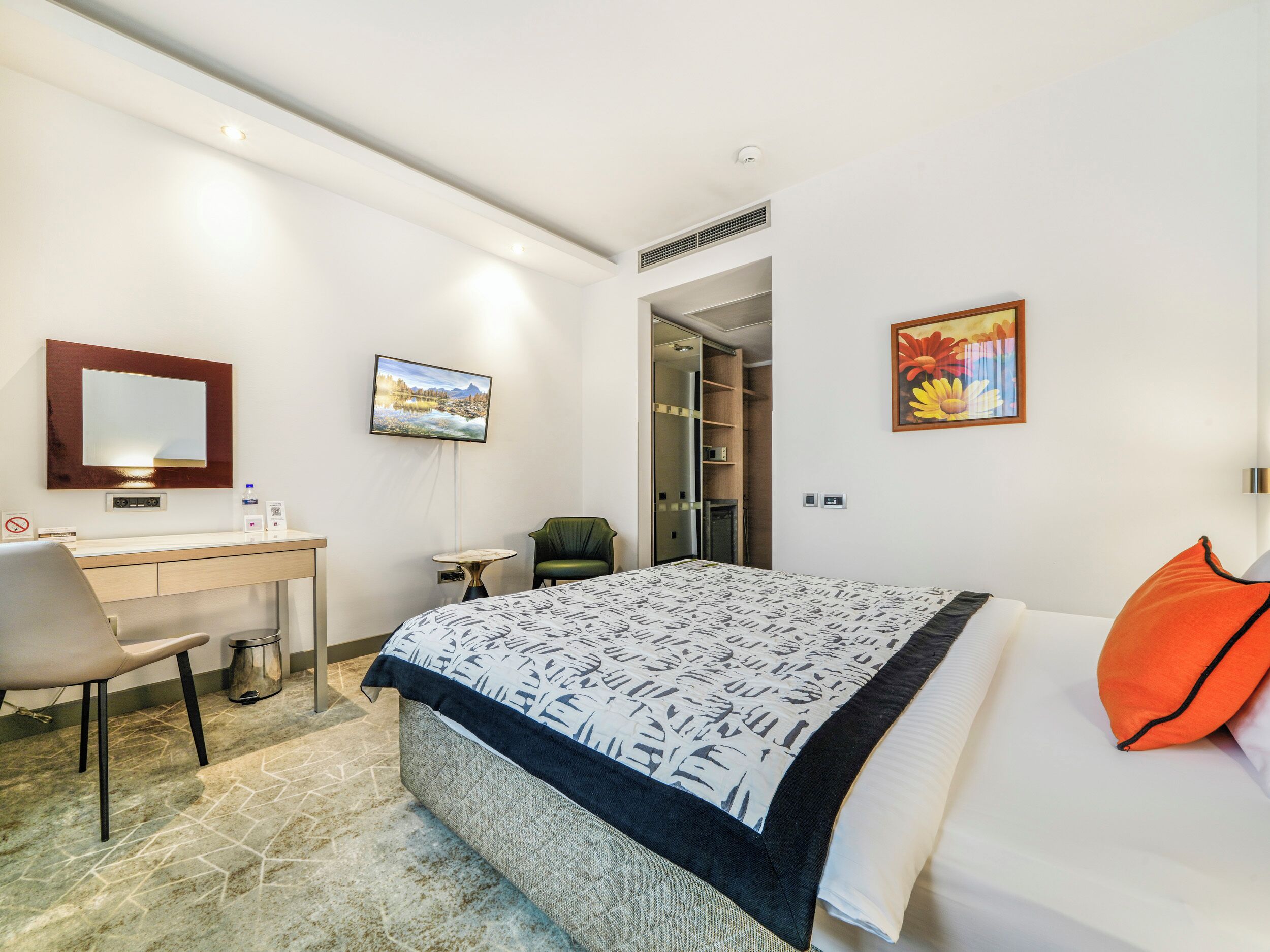 Habitación de hotel con cama, escritorio, silla, una mesita, espejo, cuadro, armario y puerta que da a un baño.