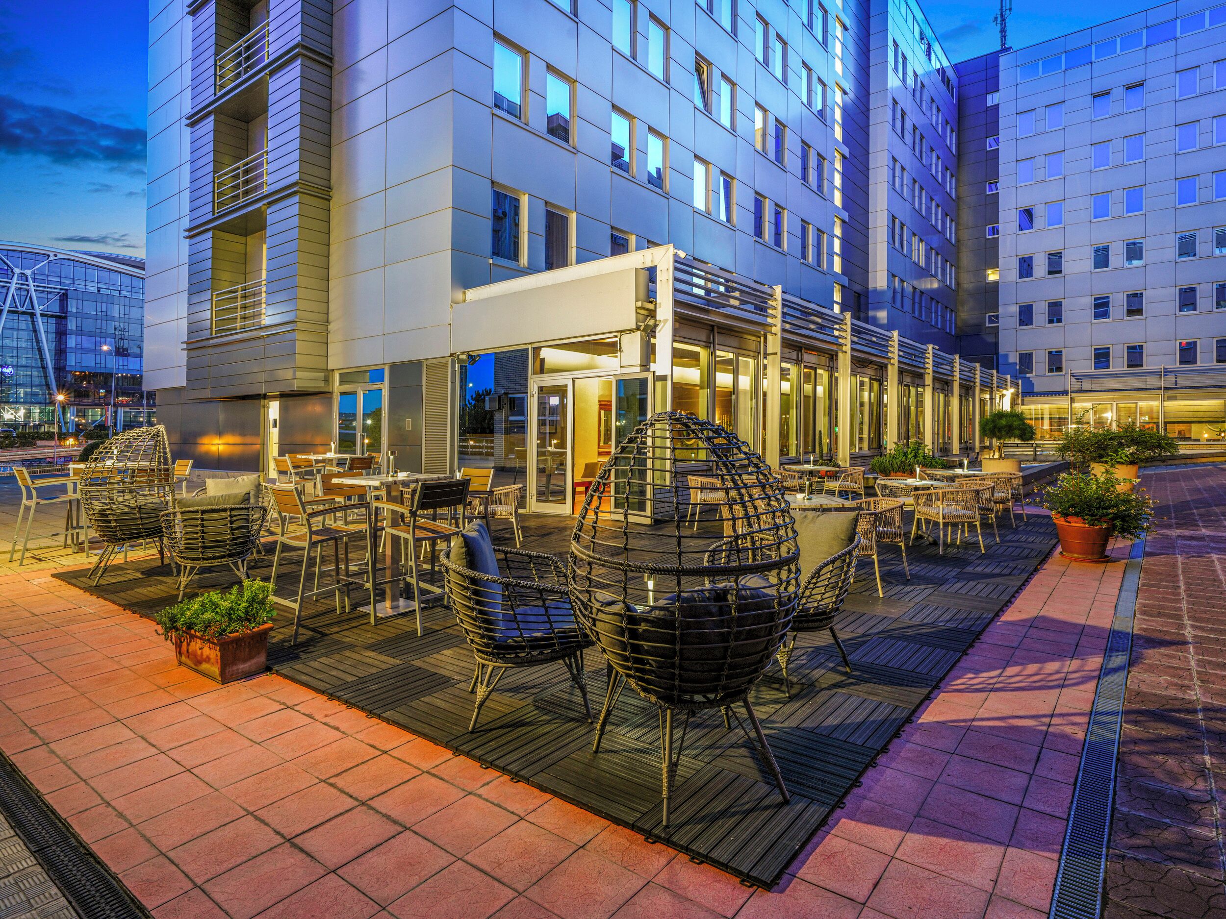 An outdoor seating area with wicker chairs and tables, a building with multiple floors and large windows, and a paved walkway.