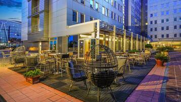 An outdoor seating area with wicker chairs and tables, a building with multiple floors and large windows, and a paved walkway.
