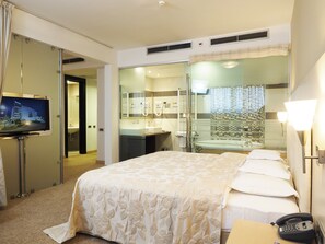 A hotel room with a bed, a TV, a bathroom with a shower, and a desk with a phone.