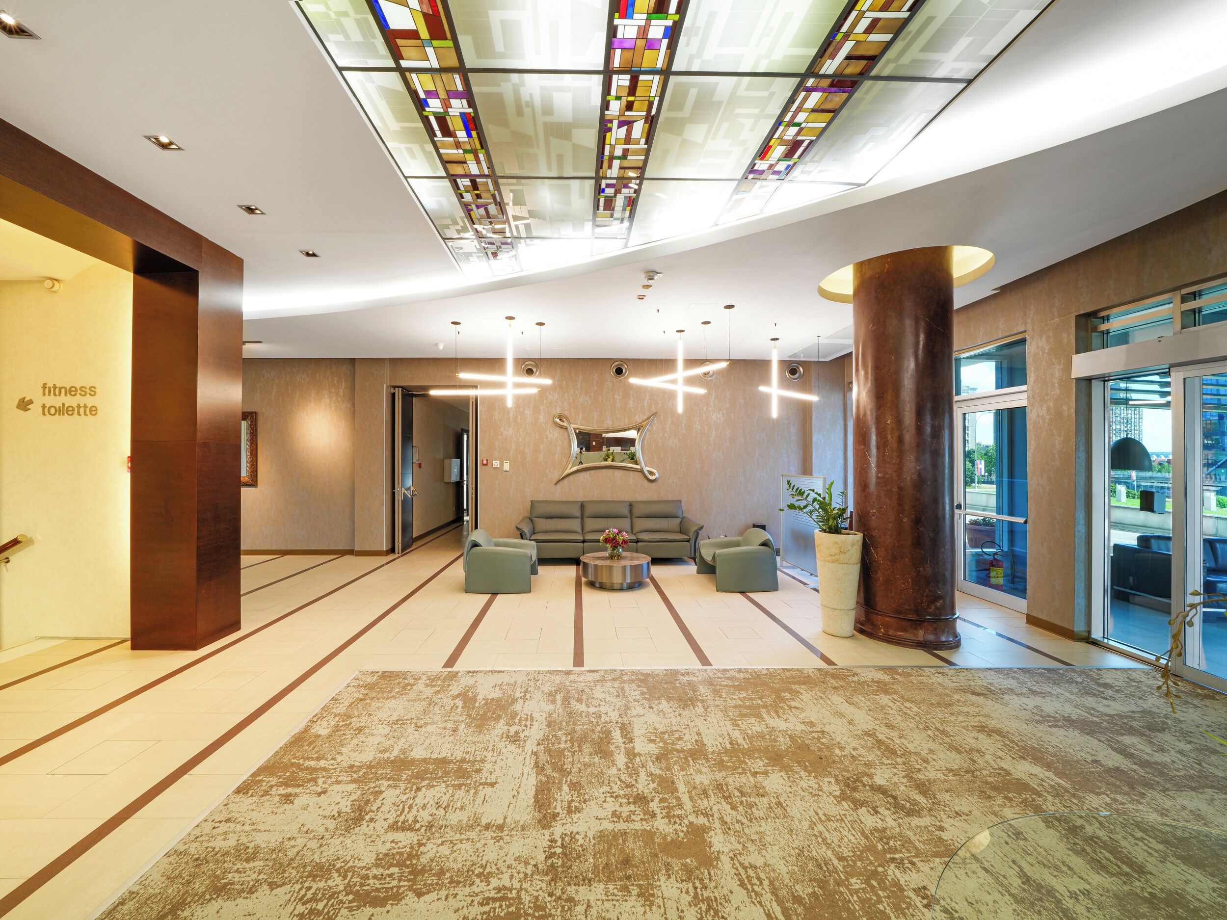 A spacious lobby with a modern design, featuring a seating area with sofas and a coffee table, a large decorative mirror, and a glass partition with a view.