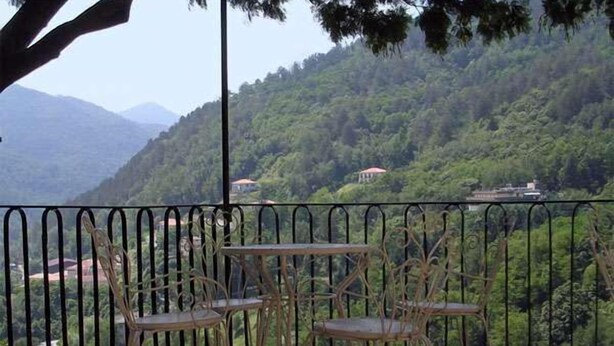A terrace with metal railings, two tables, and four chairs. The terrace overlooks a lush green hillside with a few buildings in the distance. - Agriturismo Casa del Bosco (Borzonasca)