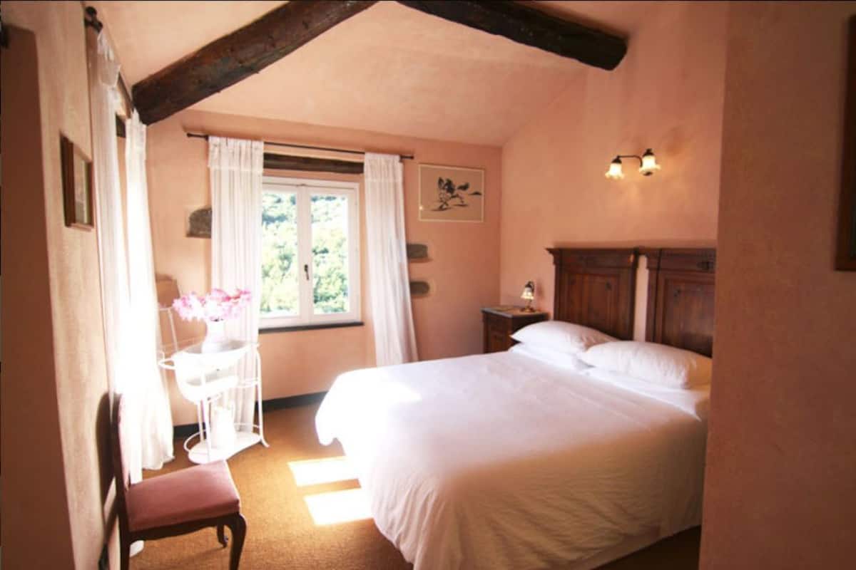 A bedroom with a large bed, a bedside table, a chair, a window with curtains, and a wall-mounted light fixture.