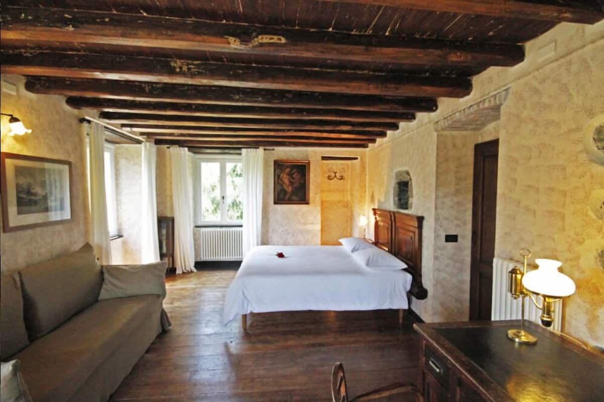 A bedroom with a wooden ceiling, a bed with white linens, a wooden nightstand, a lamp, and a window with curtains.