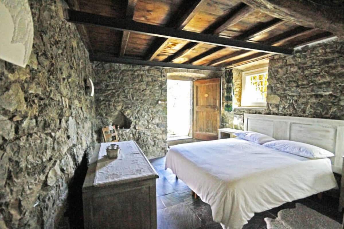 A stone-walled bedroom with a wooden ceiling, a bed, a nightstand, and a window with a view.