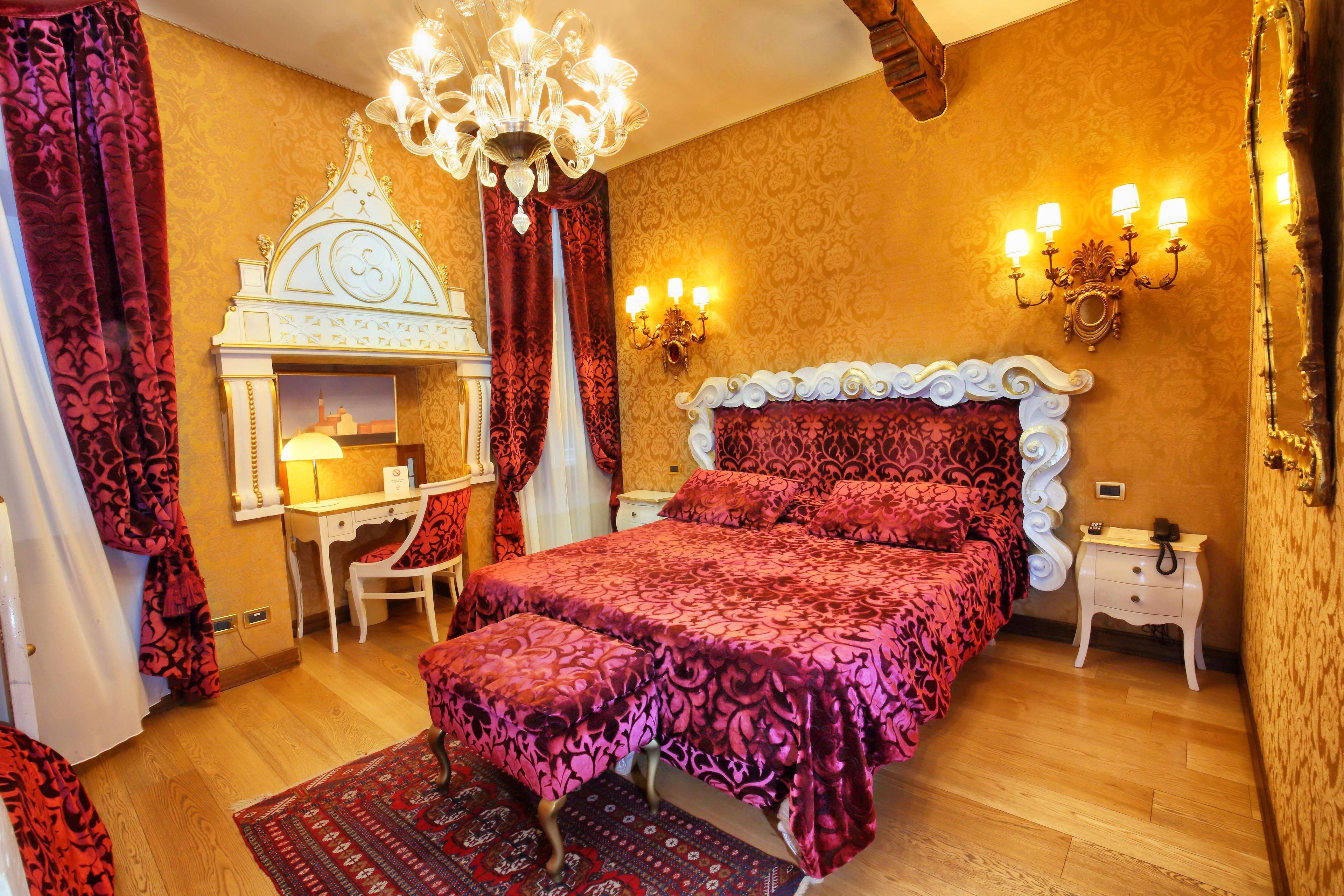 A bedroom with a large bed, a desk, and a chair. There is a chandelier, wall sconces, and a patterned rug.