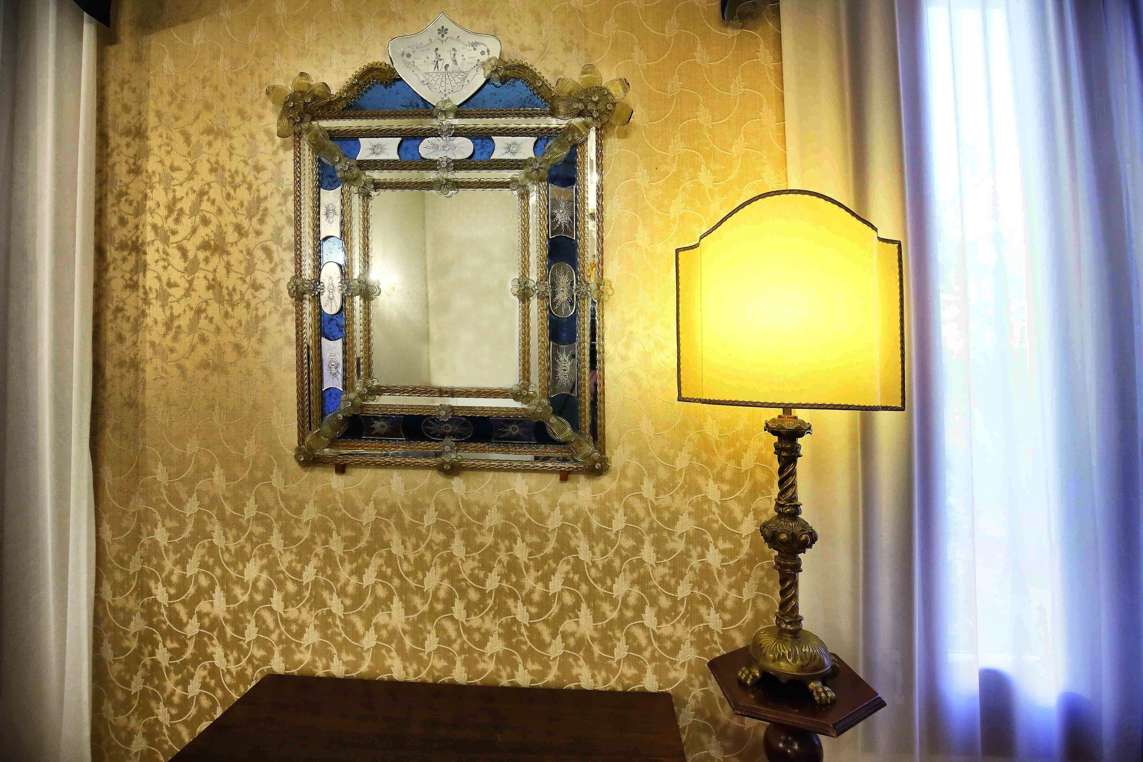 A room with a patterned wallpaper, a mirror with a decorative frame, and a table lamp.