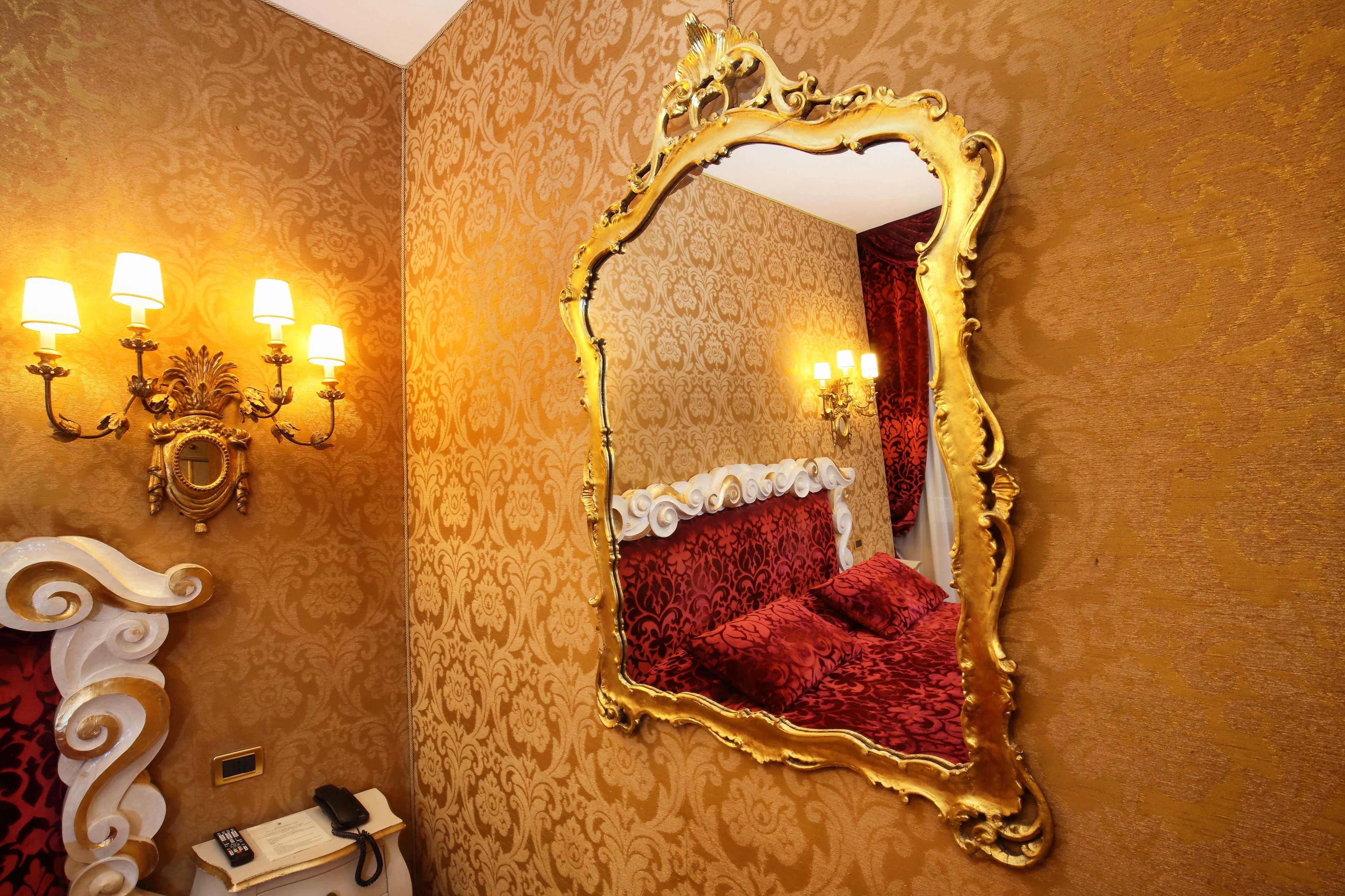 A room with a large ornate mirror, a red upholstered bed, and wall-mounted sconces.