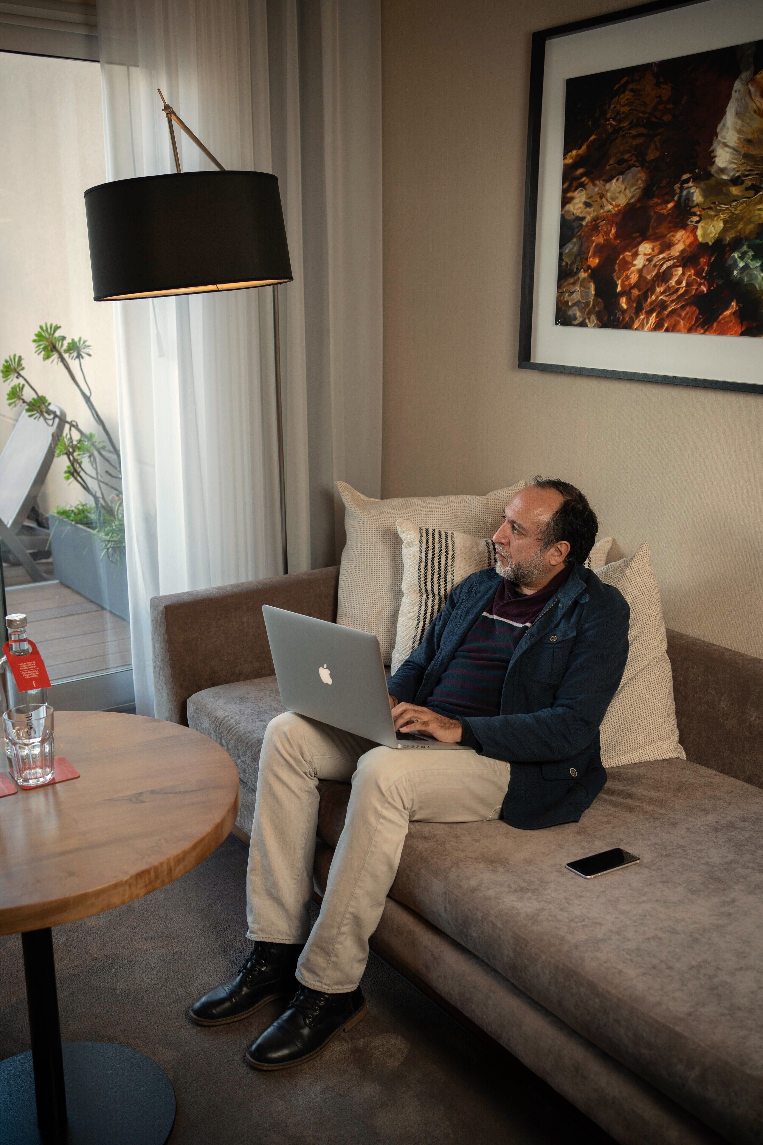 Een man zittend op een bank met een laptop, een tafel met een fles en een glas, en een lamp.
