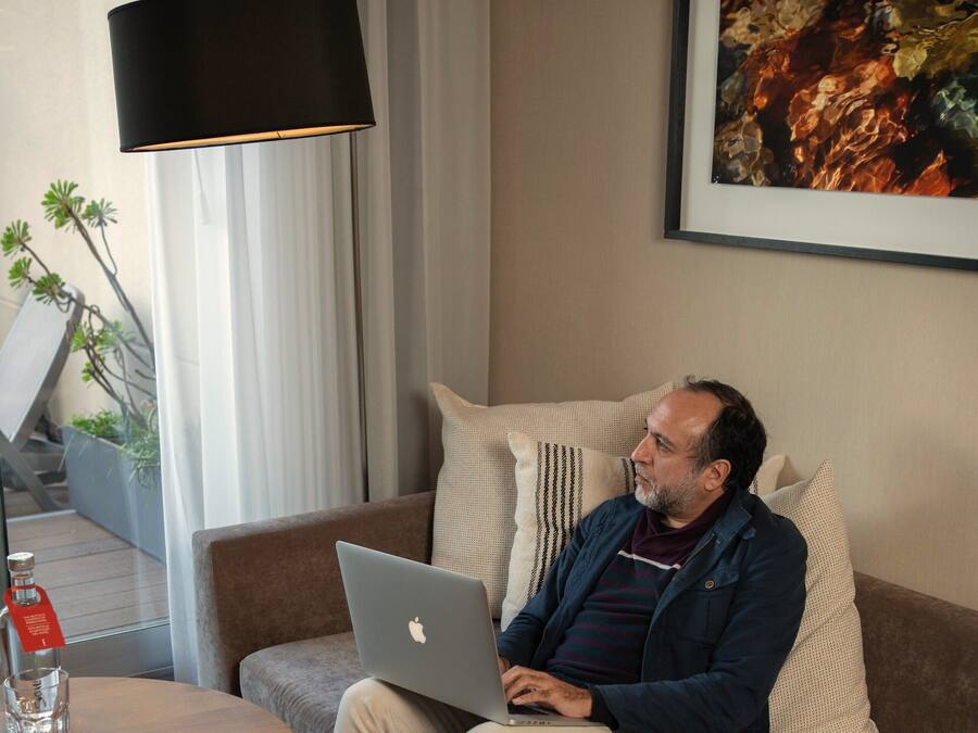 Een man zittend op een bank met een laptop, een tafel met een fles en een glas, en een lamp.
