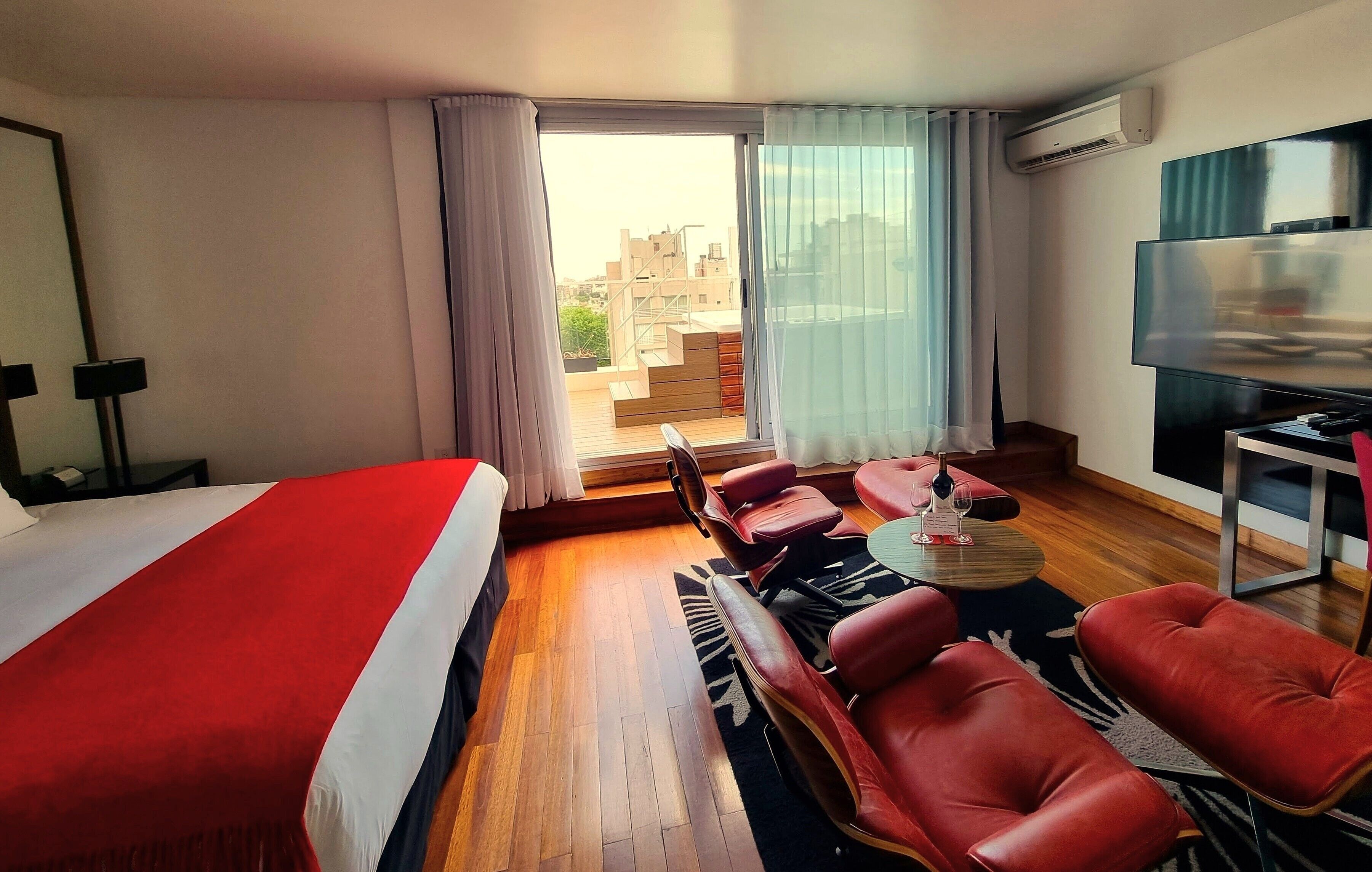Habitación de hotel con una cama grande, televisión, balcón con vistas a edificios y sillones de cuero rojo.