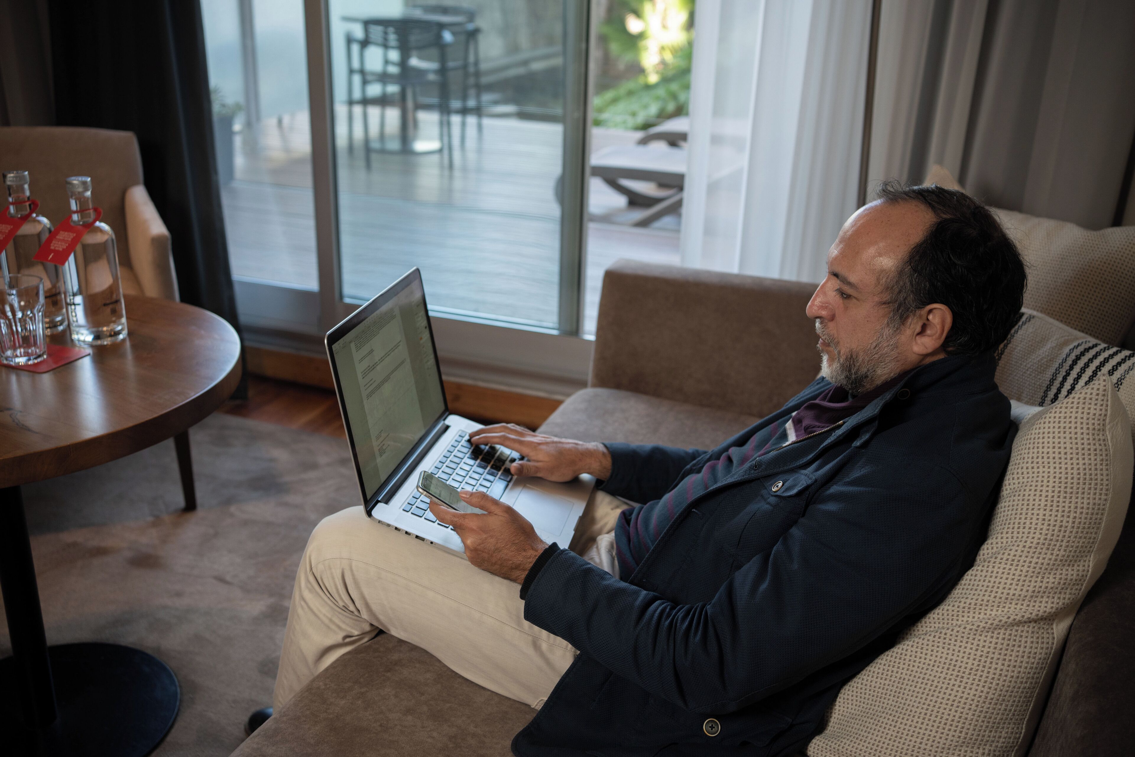 Ein Mann sitzt auf einem Sofa und benutzt einen Laptop. Im Vordergrund steht ein Tisch mit Flaschen und einer Speisekarte.