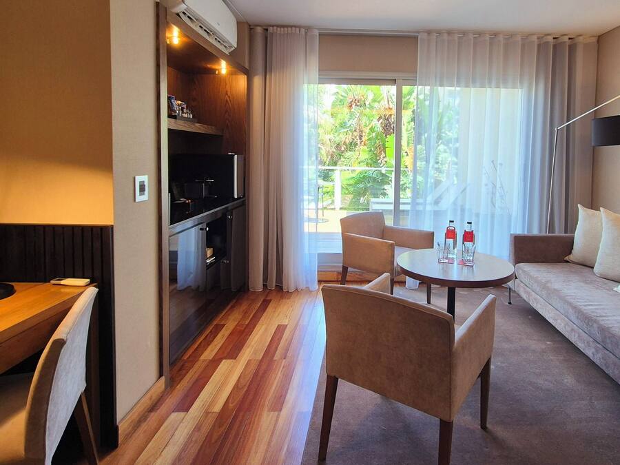 A hotel room with a dining area, a sofa, and a view of greenery outside.