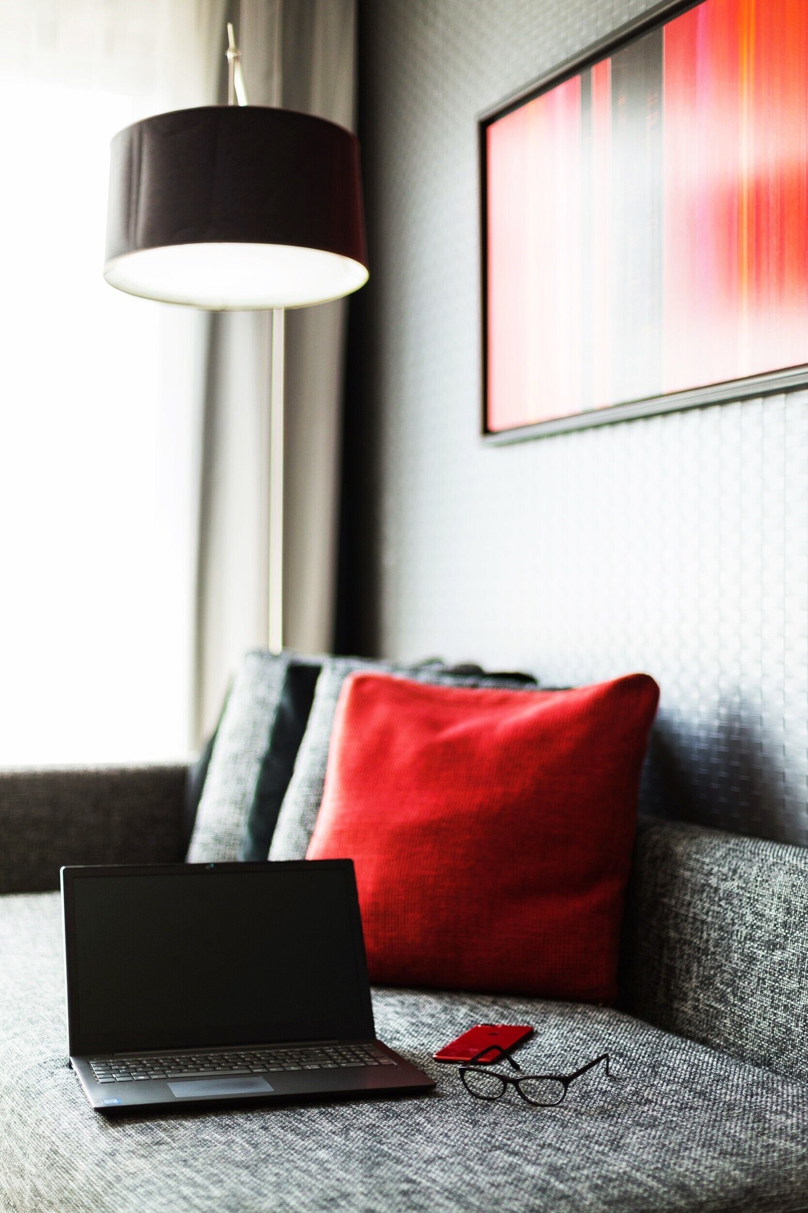 Una laptop sobre un sofá con textura, un cojín rojo, un par de gafas y una lámpara colgante.