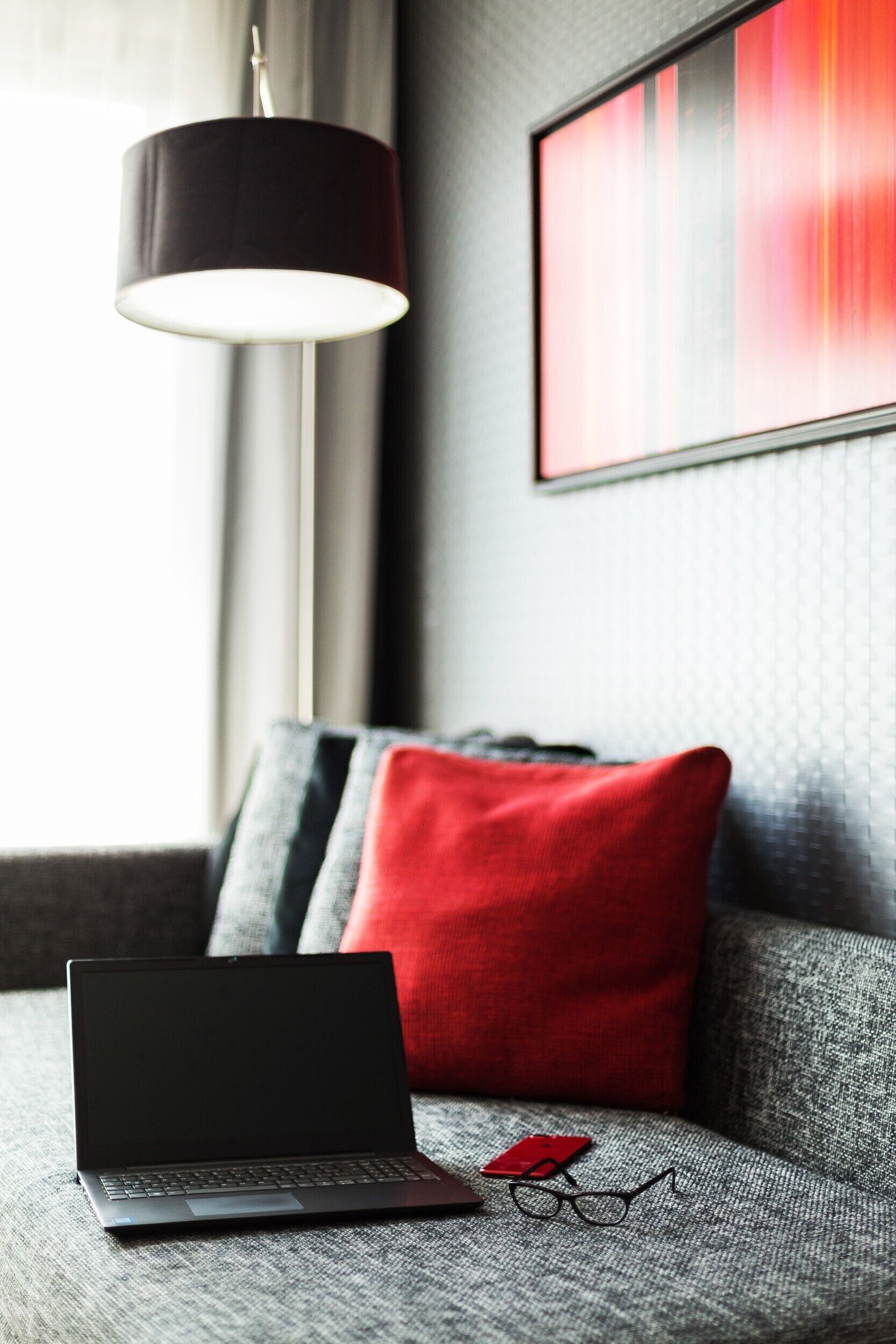 A laptop on a gray sofa with a red pillow, a black lamp, and a framed abstract painting on the wall.