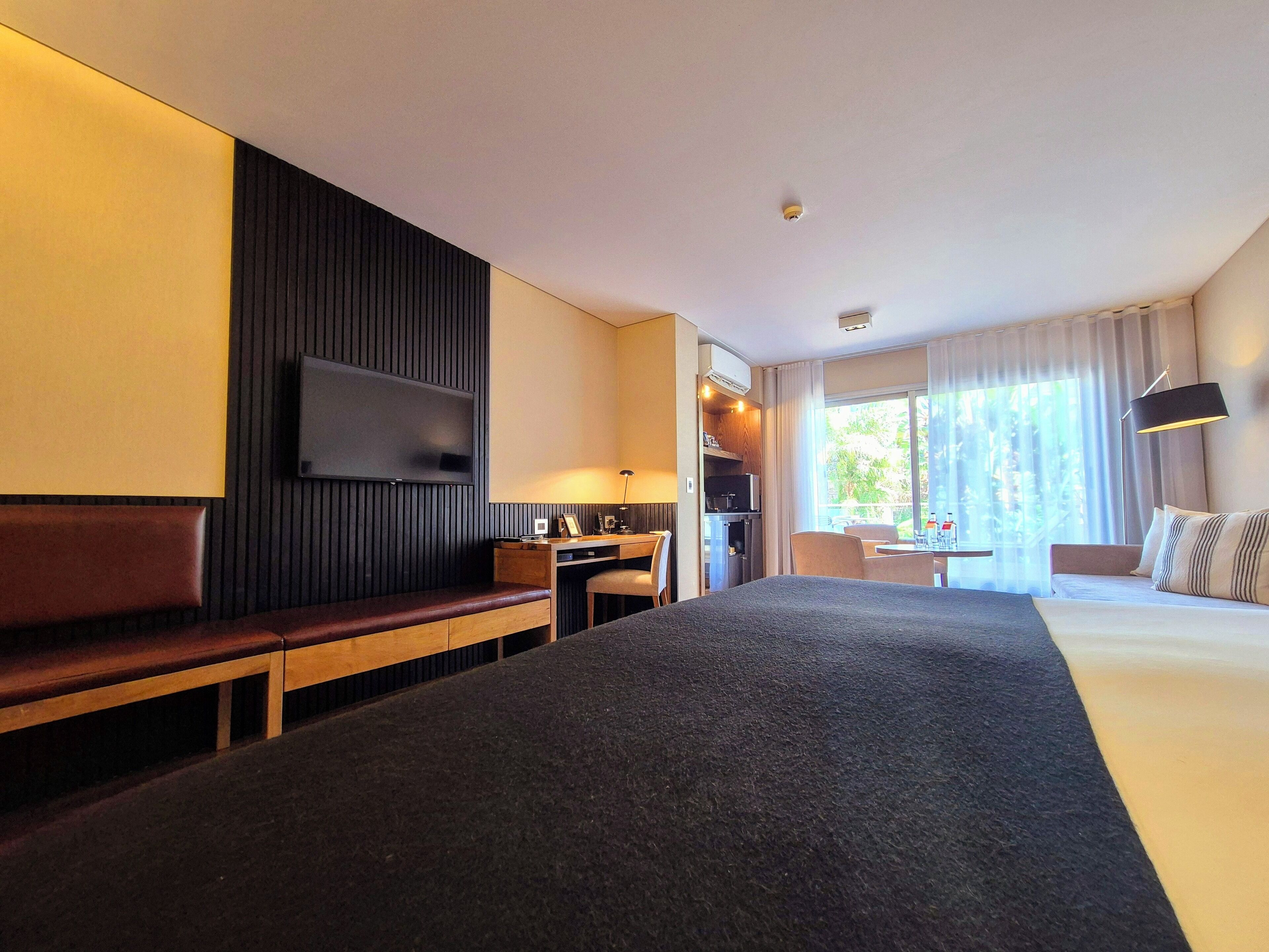 Habitación de hotel con una cama grande, un escritorio con silla, un televisor montado en la pared y una ventana con cortinas.