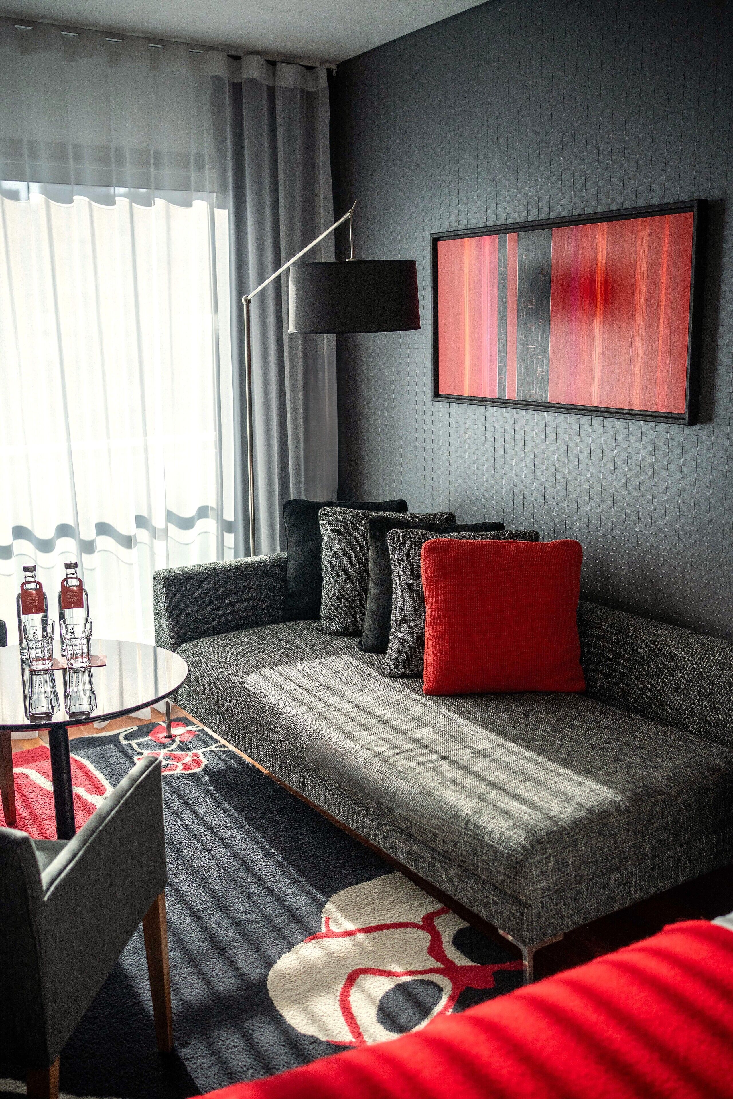 Una sala de estar moderna con un sofá gris, cojines rojos y negros, una lámpara de pie negra y un cuadro abstracto grande en la pared.