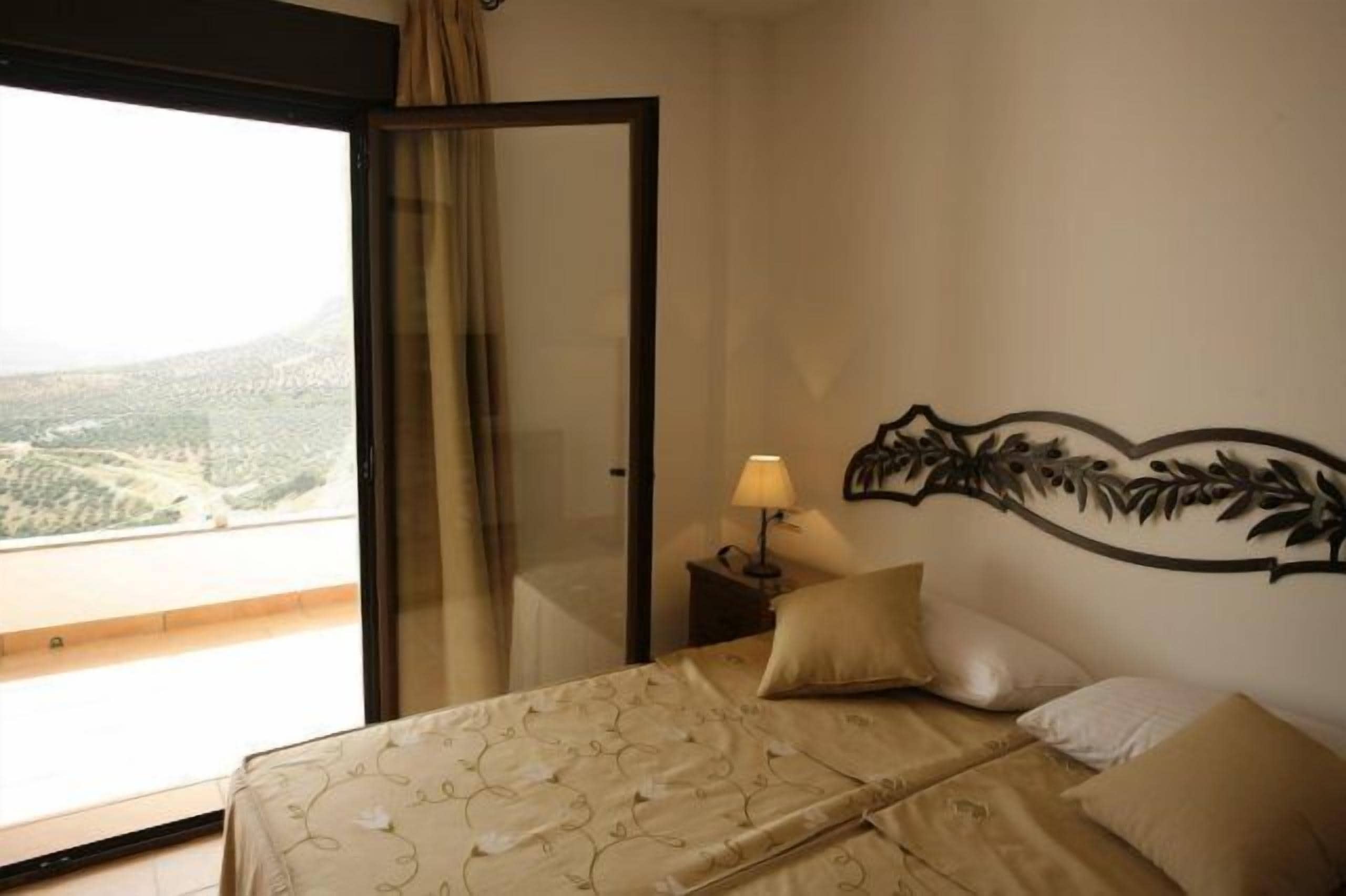 Habitación de hotel con cama, mesita de noche con lámpara, un adorno en la pared y un ventanal con vistas a un paisaje montañoso.