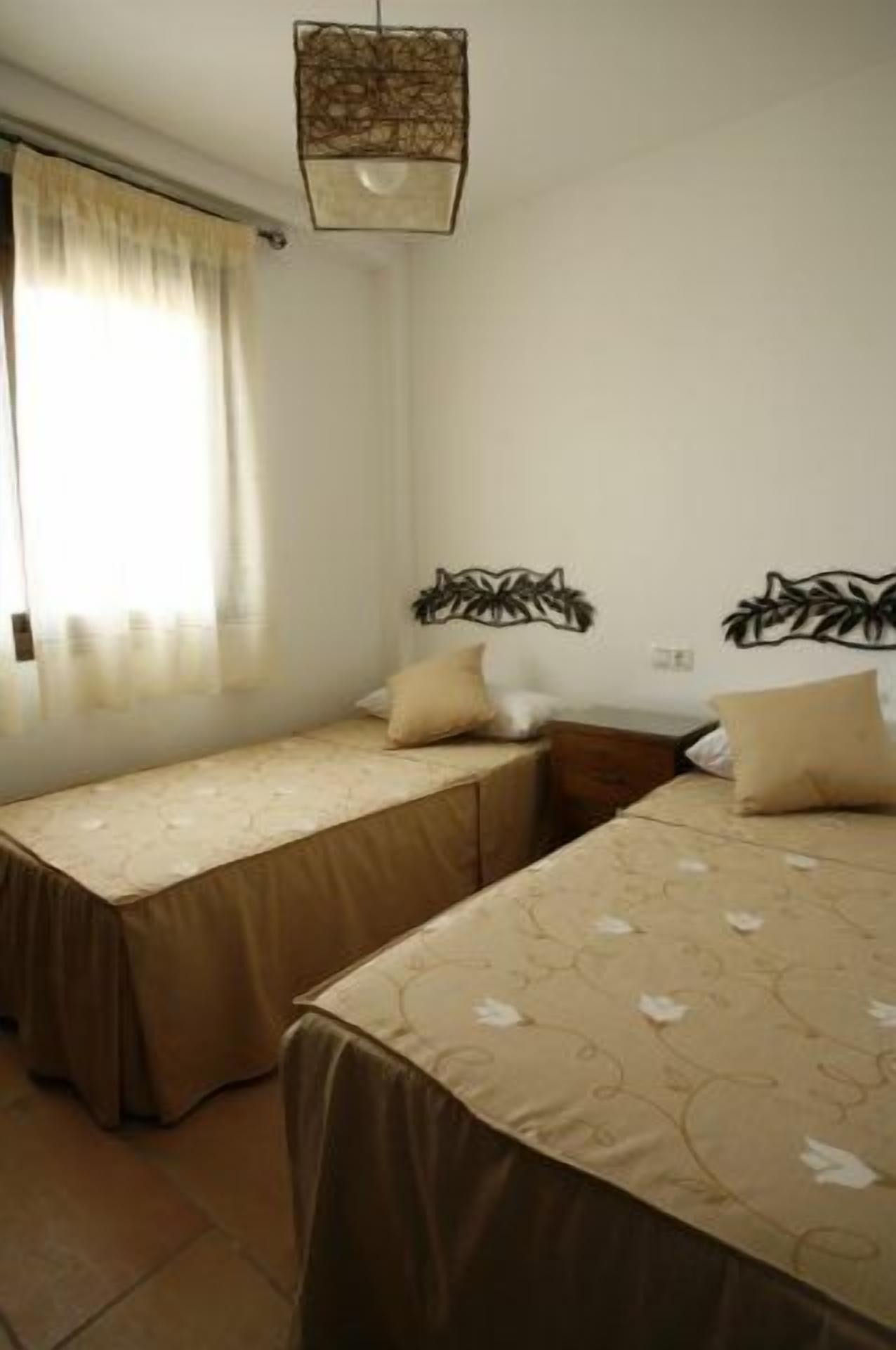 Habitación con dos camas, cada una con cubrecama marrón y una sola almohada. Se ve una ventana con cortinas translúcidas, una lámpara fijada a la pared y una mesita con una silla.