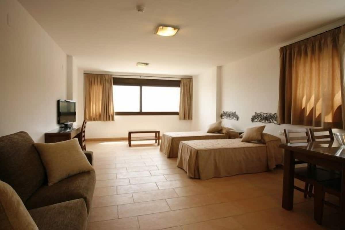 A hotel room with a sofa, two single beds, a TV, a small table, and a window with curtains.
