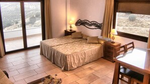 Ein Schlafzimmer mit einem Bett, einem Tisch und einem Stuhl. Vor den Fenstern hängen Vorhänge und auf dem Nachttisch steht eine Lampe.