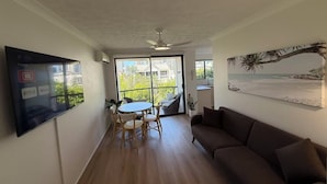 A living room with a TV, a couch, a dining table with chairs, and a balcony with a view of buildings and greenery. - Chevron Palms Holiday Apartments by Gold Coast Premium (Surfers Paradise)
