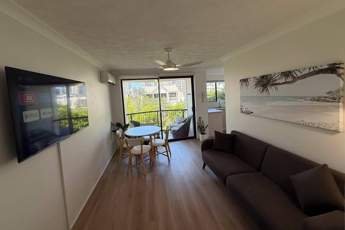 Una sala de estar con televisor, sofá, mesa de comedor con sillas y balcón con vista a edificios y vegetación.