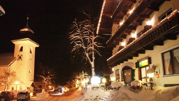 A snow-covered street with a church and a building with a balcony. - Gasthof Goldener Stern (Sankt Koloman)