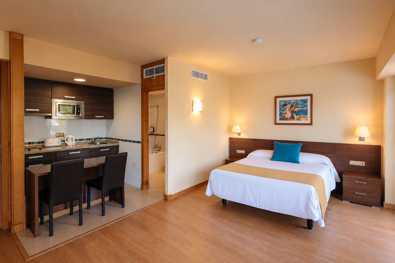 A hotel room with a bed, bedside table, lamp, and a kitchenette with a microwave.