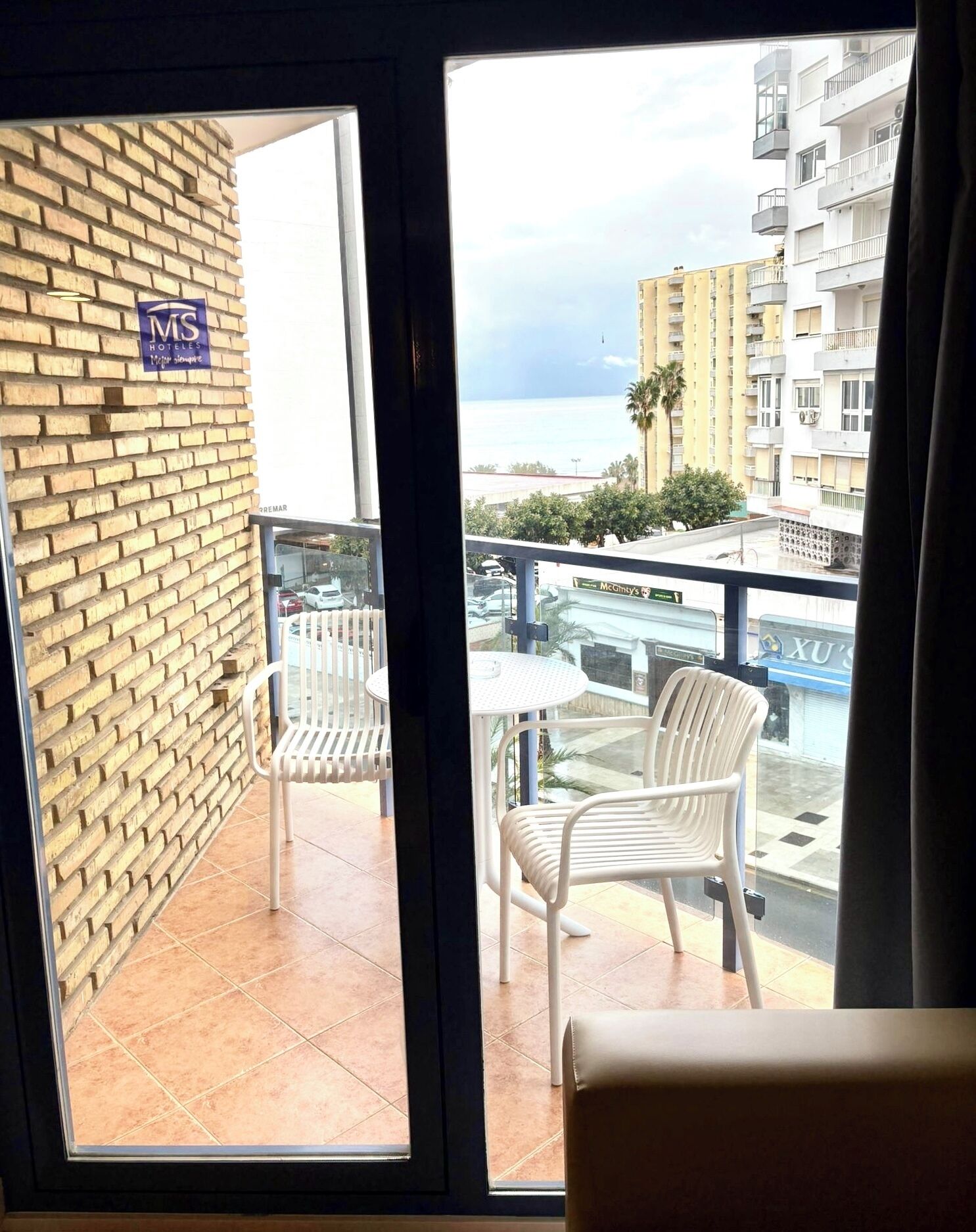 Un balcón con dos sillas blancas, un muro de ladrillo y vista al mar y edificios.