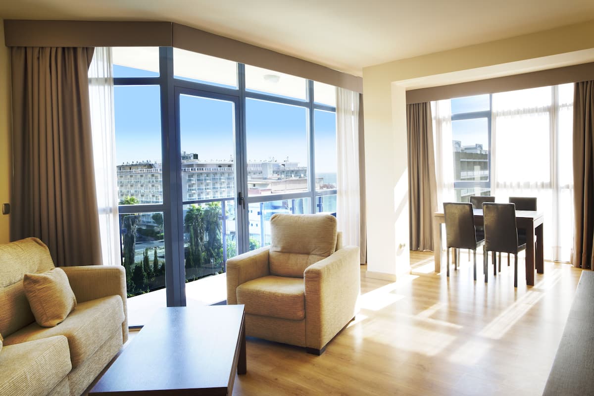Ampla sala de estar com sofá, poltrona, mesa de jantar e cadeiras. Janelas grandes oferecem vista para o horizonte urbano.