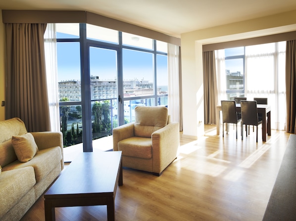A spacious living area with a sofa, armchair, dining table, and chairs. Large windows offer a view of the cityscape.