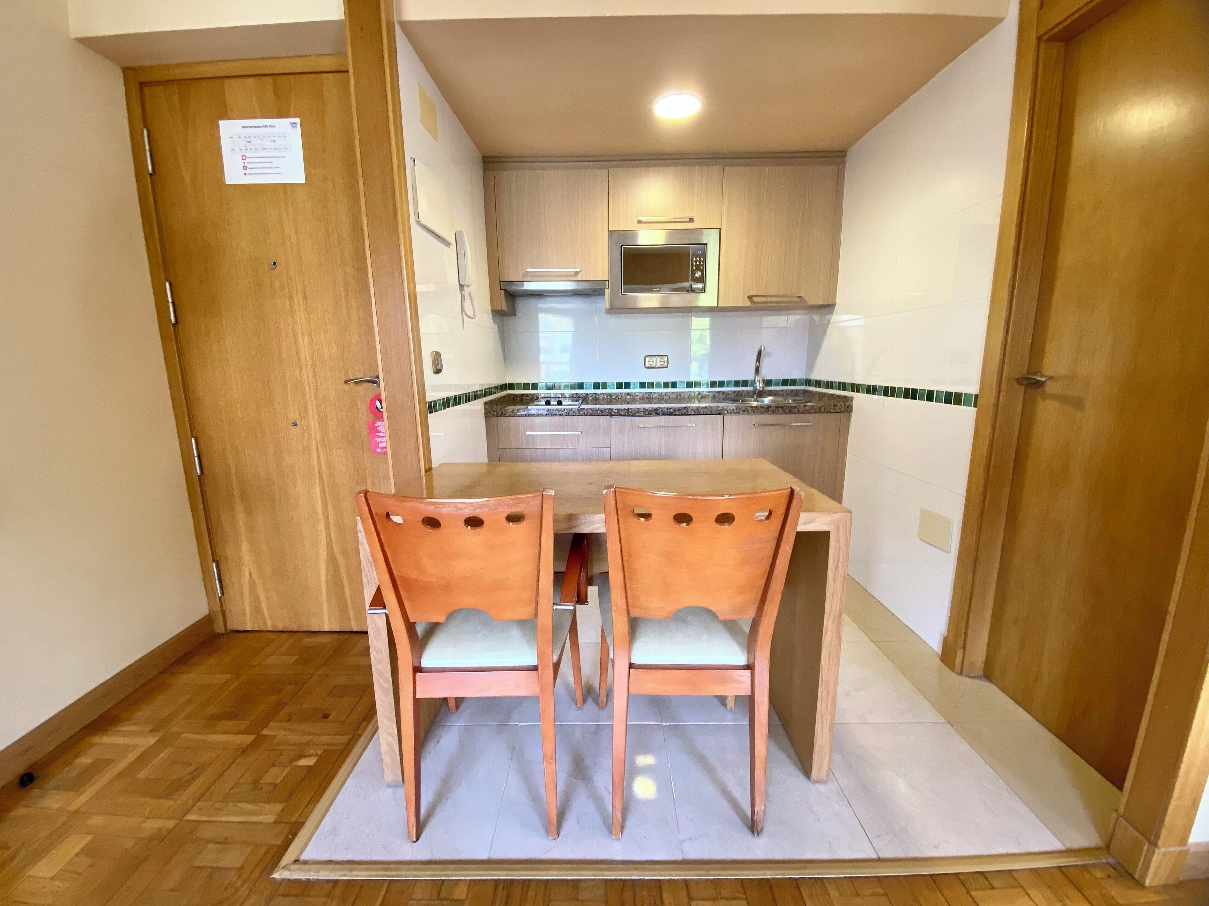 A compact kitchen with wooden cabinets, a microwave, and a sink. There are two wooden chairs and a small table.