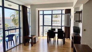 Een ruime woonkamer met houten vloer, een eettafel met stoelen, een tv op een dressoir en grote ramen met uitzicht op de stad.