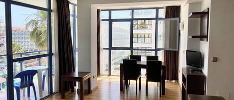 Amplia sala de estar con piso de madera, una mesa de comedor con sillas, un televisor sobre un mueble bajo y ventanas grandes con vistas a la ciudad.