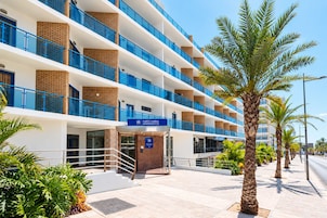 A multi-story hotel with a modern design, featuring balconies, a central entrance, and a clear sky.