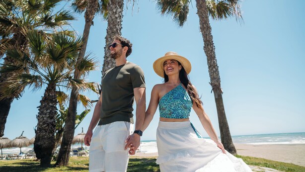 Een stel dat hand in hand over het strand loopt, met palmbomen en een heldere lucht.