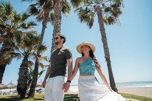 Una pareja tomados de la mano en una playa con palmeras y cielo despejado.