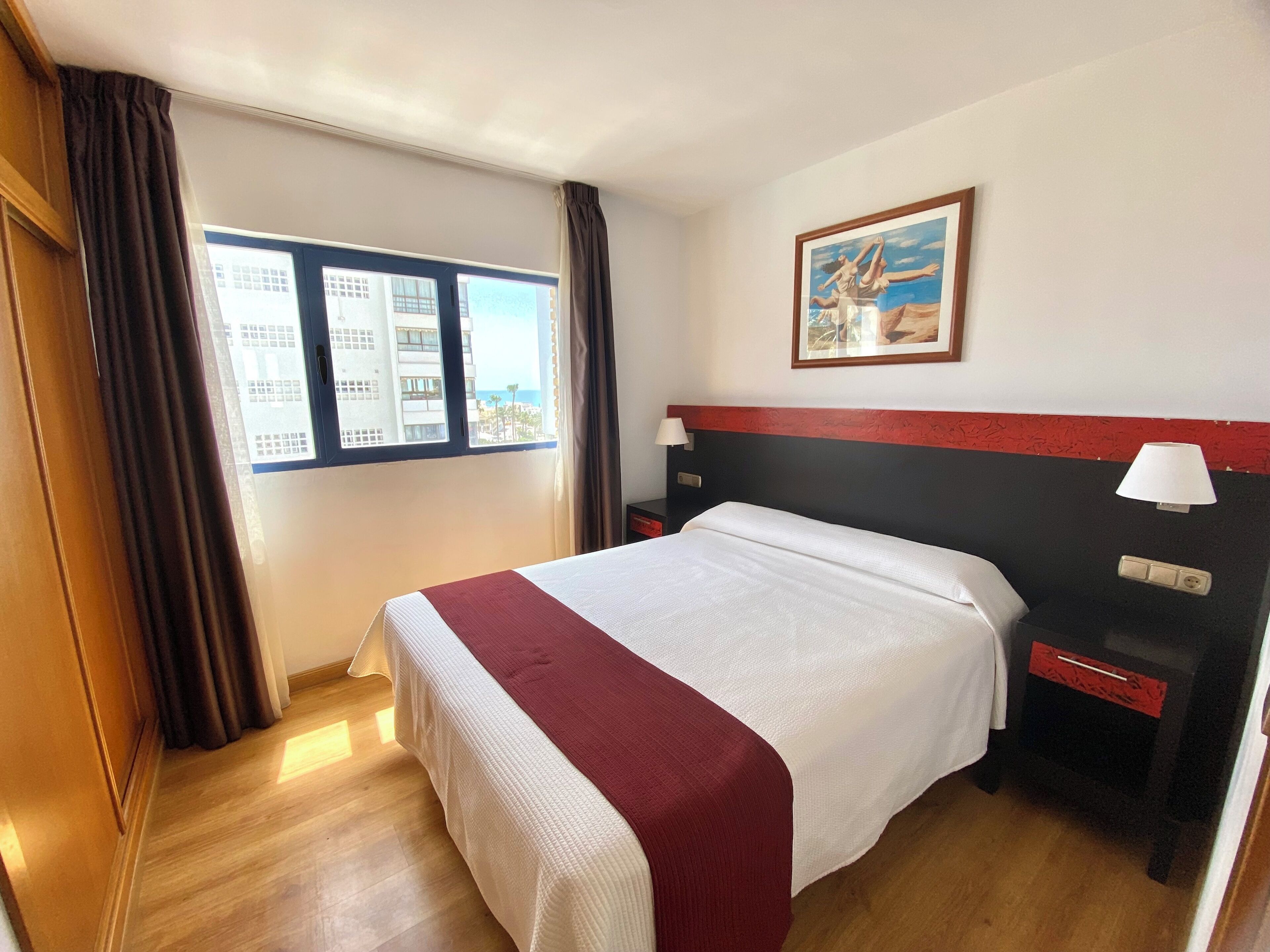 Una camera d'albergo con un letto, un comodino, una lampada, una finestra con tende e un quadro appeso al muro.