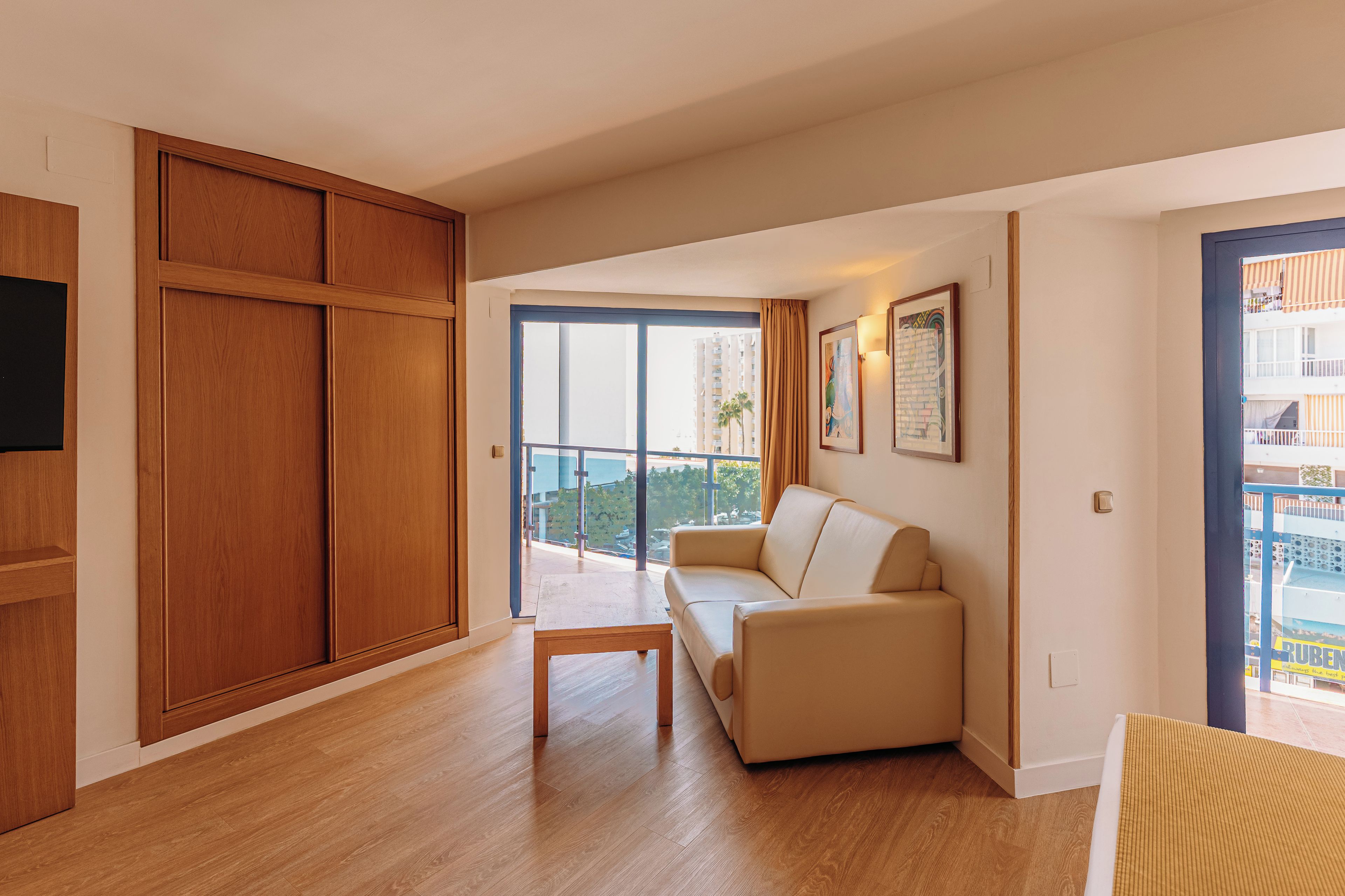 A modern living room with a wooden floor, a beige sofa, a small wooden table, and a large window with a view of buildings and greenery.