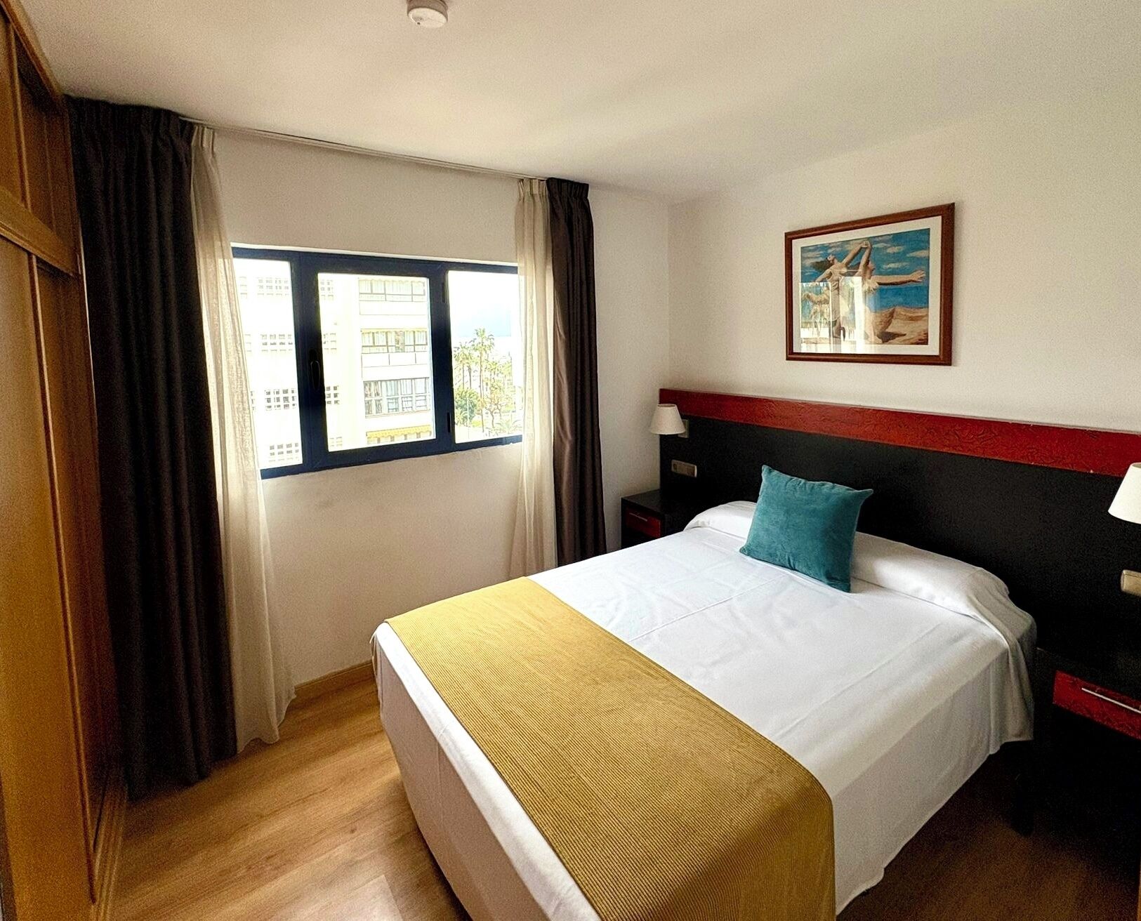 A hotel room with a bed, a window with curtains, a painting on the wall, and a wooden floor.