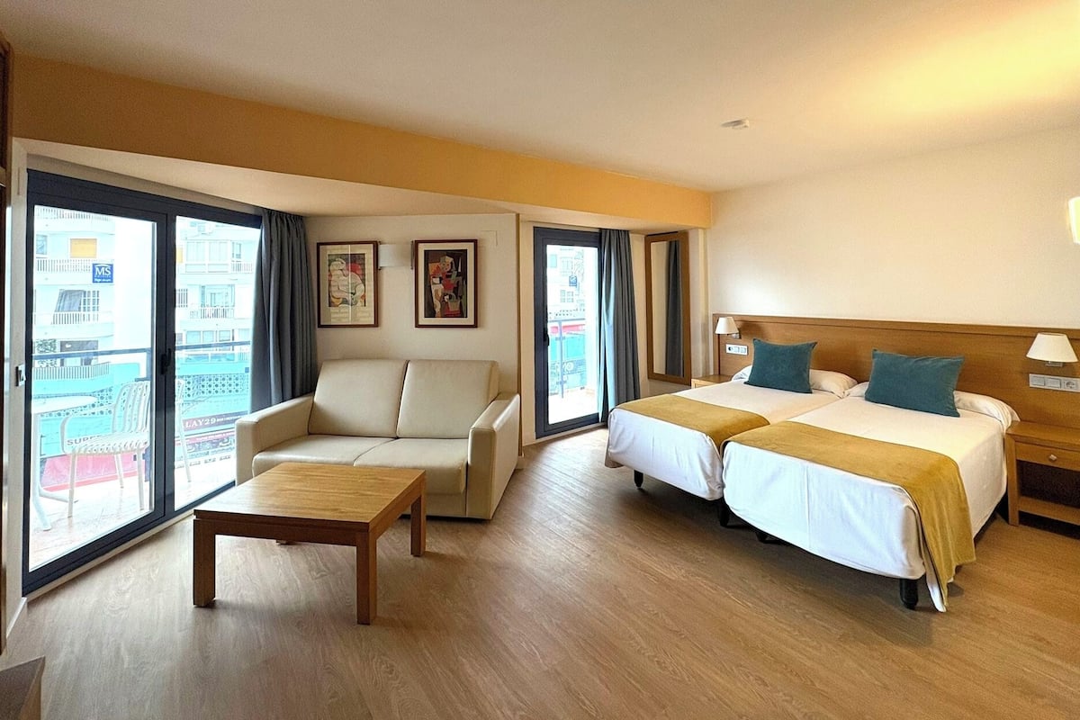 Quarto de hotel com sofá, duas camas, uma mesa e grandes janelas com vista para a cidade.