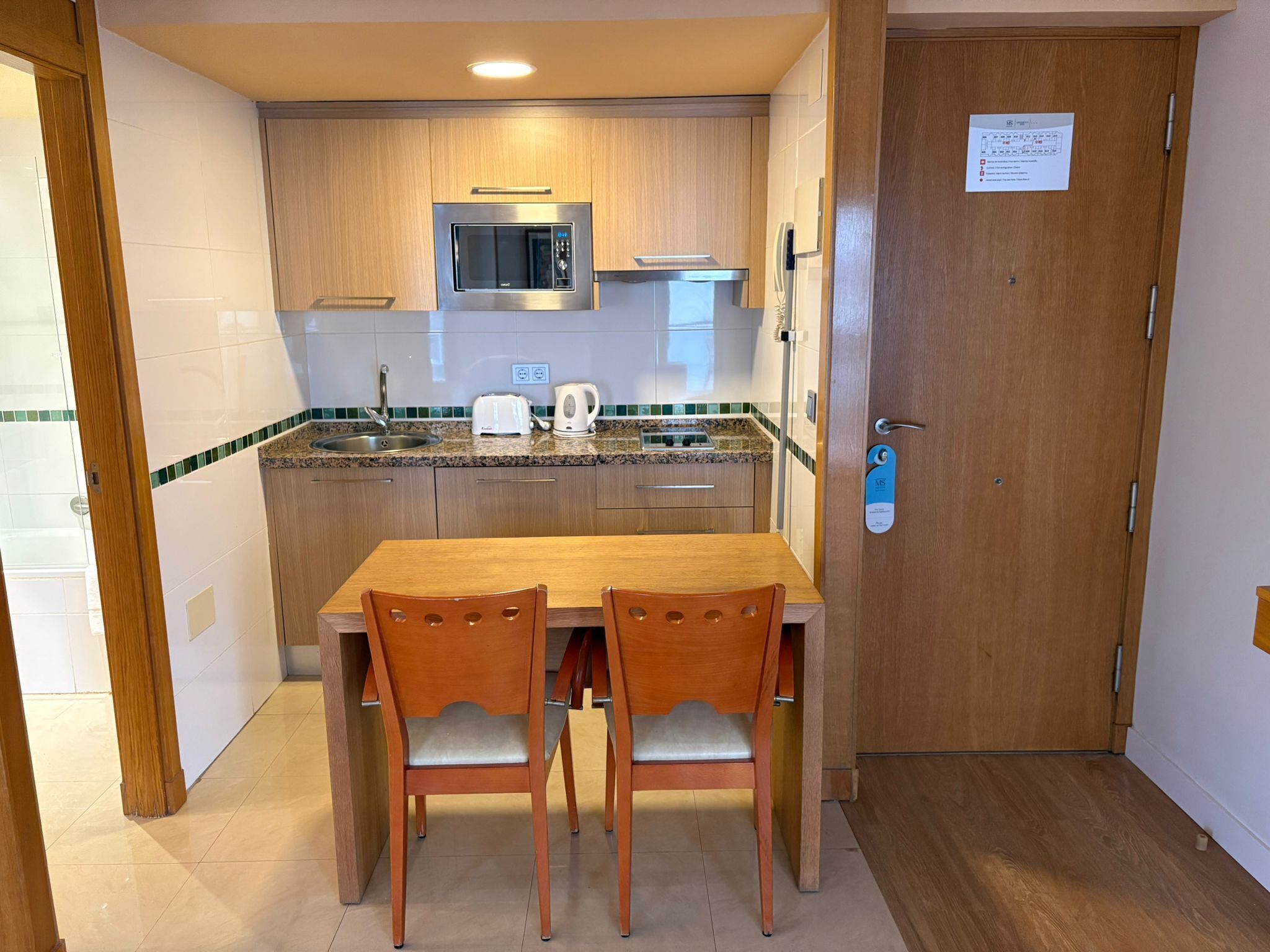 A compact kitchen with a microwave, toaster, and kettle. There is a wooden table with two chairs and a door with a keycard lock.