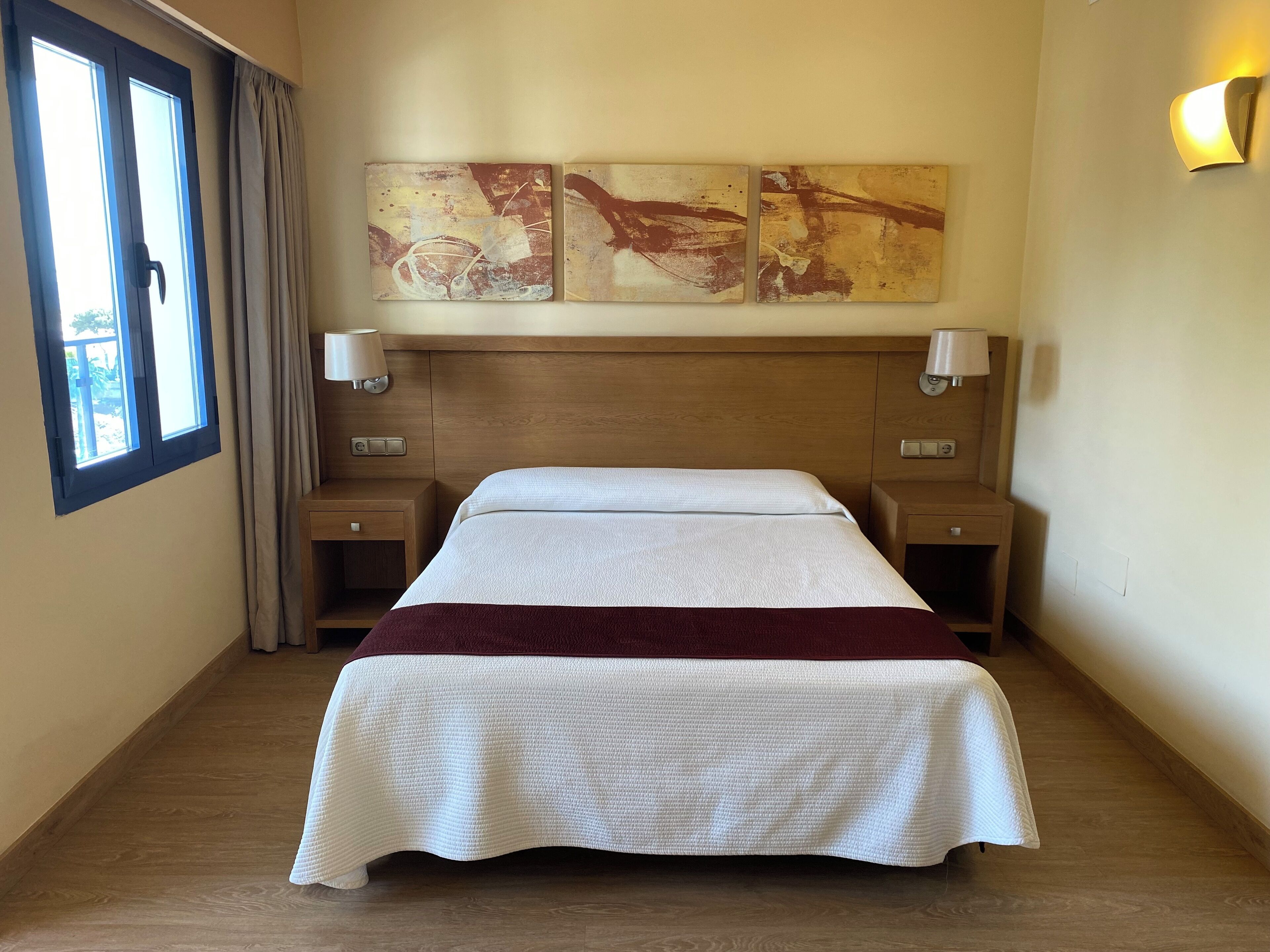 Ein Hotelzimmer mit Doppelbett, zwei Nachttischen, einem Fenster mit Vorhängen und zwei Wandlampen.