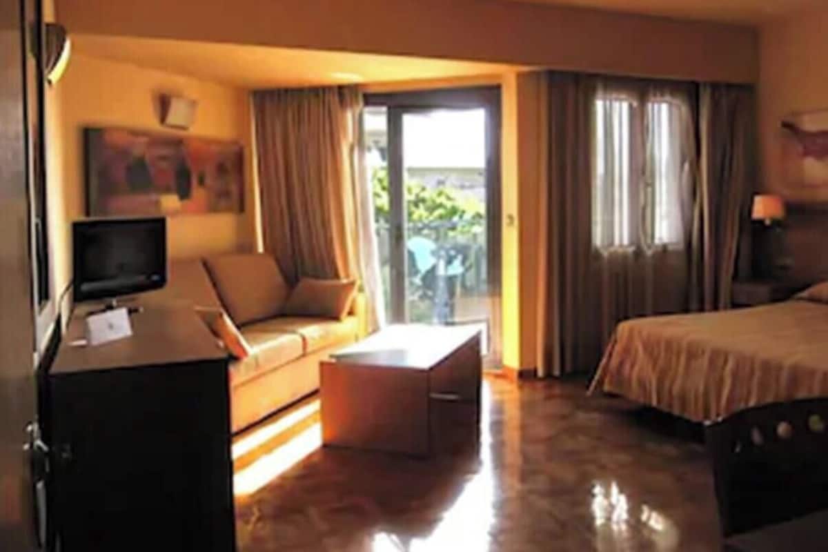 Quarto de hotel com cama, sofá, mesa de centro, televisão e janela com cortinas.
