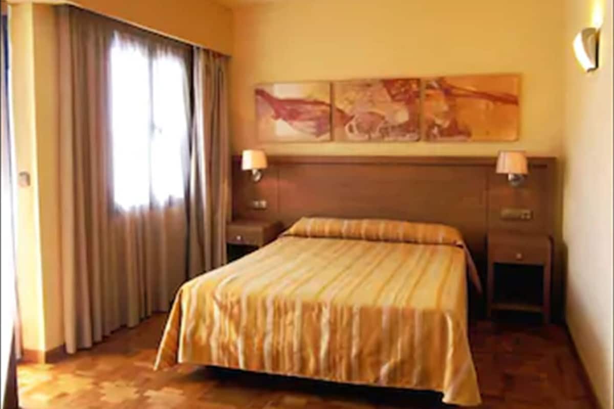 Quarto de hotel com cama grande, duas mesinhas de cabeceira, cabeceira e janela com cortinas.